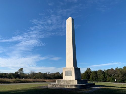 A tall, rectangular monument stands on a grassy field under a blue sky with scattered clouds.