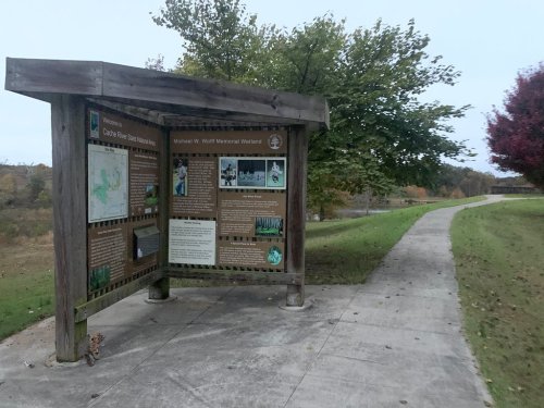 Information kiosk about Michael W. Wolf Memorial Wetland, with a path and trees in the background.