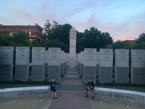 Two children sit on a pathway in a veterans memorial, surrounded by stone pillars commemorating World Wars I and II.
