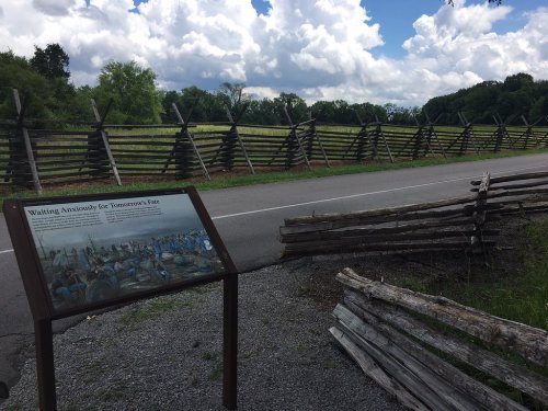 A scenic view of a wooden fence along a road, with a sign about historical events and a cloudy sky above.