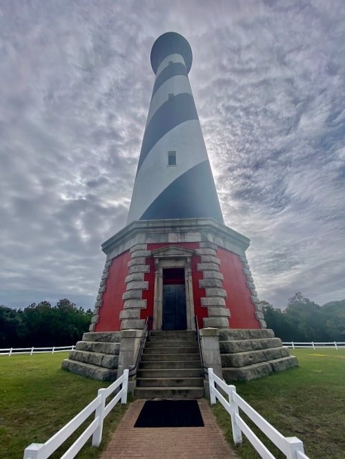 A tall striped lighthouse with a red base, surrounded by green grass and a cloudy sky. Steps lead to its entrance.