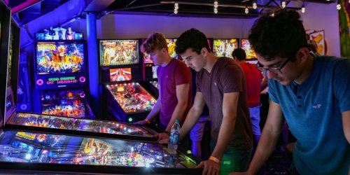 Three young men play pinball in a vibrant arcade filled with colorful machines and ambient lighting.