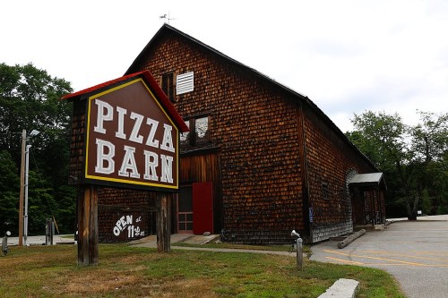 A rustic wooden building with a large sign reading "Pizza Barn," featuring a welcoming entrance and parking area.