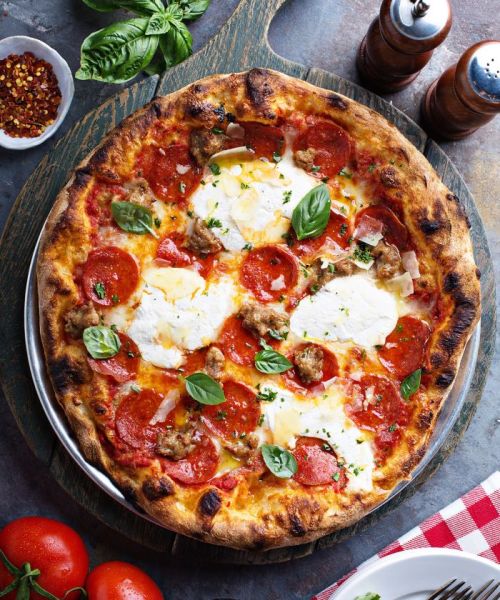 A freshly baked pizza topped with pepperoni, sausage, mozzarella, and fresh basil, served on a wooden board.