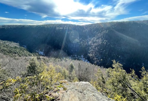 A scenic view from a rocky overlook, showcasing a valley with trees and a bright sky. Sunlight beams through the clouds.