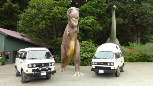 Two vintage vans parked near large dinosaur sculptures in a lush green setting.