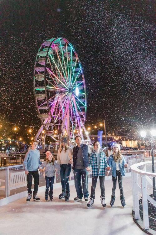 A group of six people ice skating at night, with a colorful ferris wheel and snowflakes in the background.