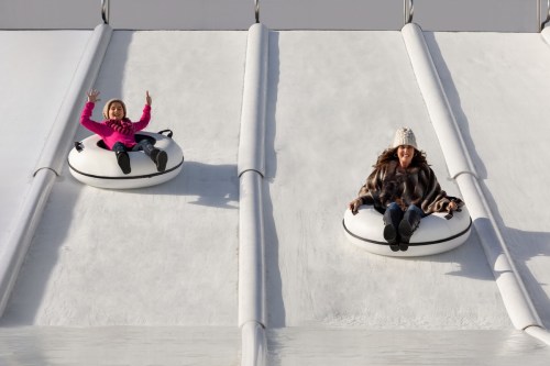 Two women on snow tubes sliding down a slope, one waving and wearing a pink jacket, the other in a fur coat and hat.