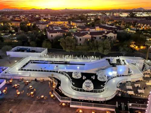 Aerial view of a rooftop venue with a winding pool, seating areas, and a vibrant sunset over the city skyline.
