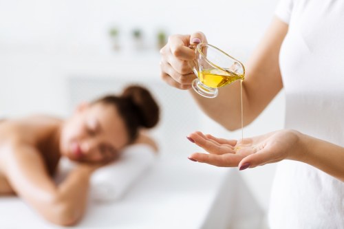 A person pours massage oil into their hand while another person relaxes on a massage table in the background.