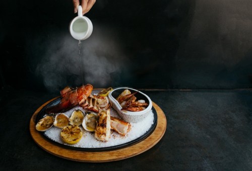 A seafood platter with grilled fish, shrimp, and charred lemons, being drizzled with sauce on a wooden board.