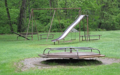 A playground with a slide, swings, and a merry-go-round, surrounded by green grass and trees.