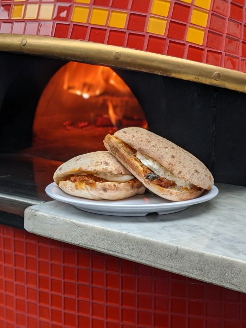 Two sandwiches on a plate in front of a brick oven, with warm colors and a cozy atmosphere.