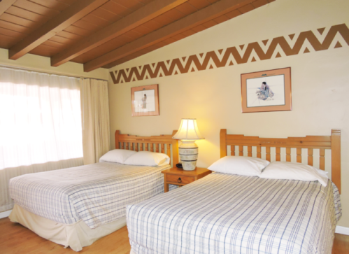 A cozy hotel room with two beds, wooden furniture, and decorative wall art. Natural light filters through a window.