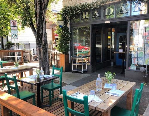 Outdoor dining area with wooden tables and green chairs, surrounded by trees, in front of a restaurant named "Radian."