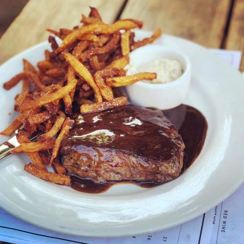 A plate featuring a juicy steak with a rich sauce, accompanied by crispy fries and a small dish of dipping sauce.