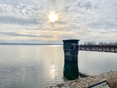 A serene lake scene with a wooden pier, a small building, and a cloudy sky reflecting on the water's surface.