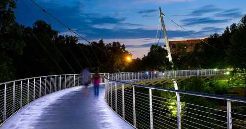 A modern pedestrian bridge illuminated at dusk, with people walking and trees in the background.