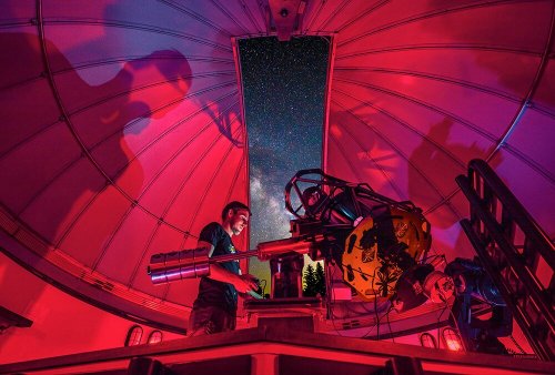 A person operates a telescope inside an observatory, with the Milky Way visible through the dome's opening.