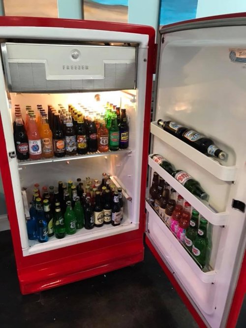 A vintage red refrigerator filled with various bottles and cans of beverages, including sodas and beers.