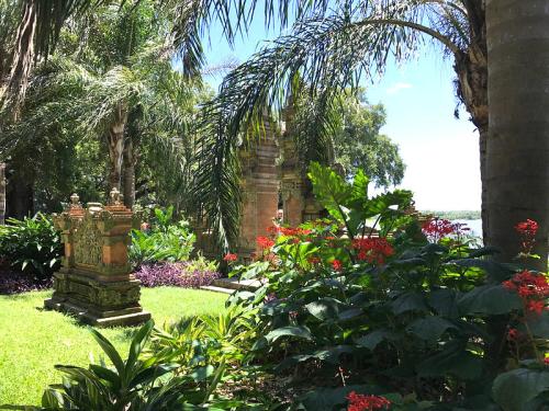 Lush garden with tropical plants, vibrant flowers, and ornate stone structures near a body of water under a clear blue sky.