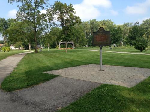 A park scene featuring a grassy area, a historical marker, and a gazebo in the background under a partly cloudy sky.