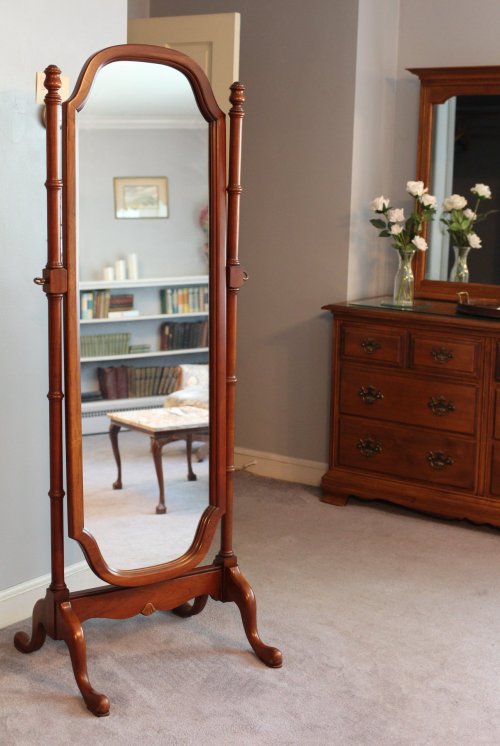 A tall, wooden standing mirror with a decorative frame, reflecting a room with a dresser and flowers.