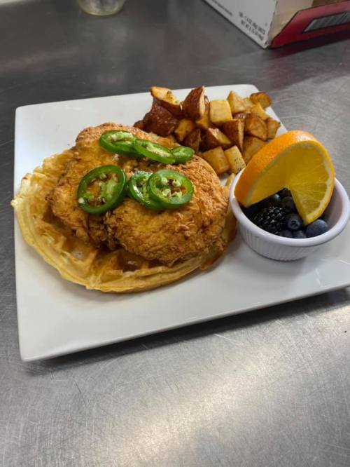 A plate featuring fried chicken on a waffle, topped with jalapeños, served with potatoes and a side of fruit.