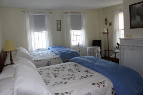 A cozy bedroom with two beds, light curtains, and a small table by the window, featuring floral bedding and a warm atmosphere.