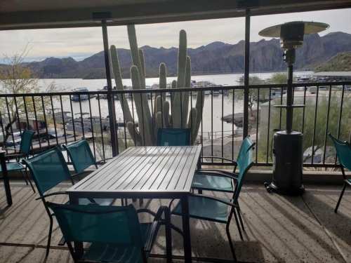 A patio with turquoise chairs and a table overlooks a marina and mountains, with a cactus in the foreground.