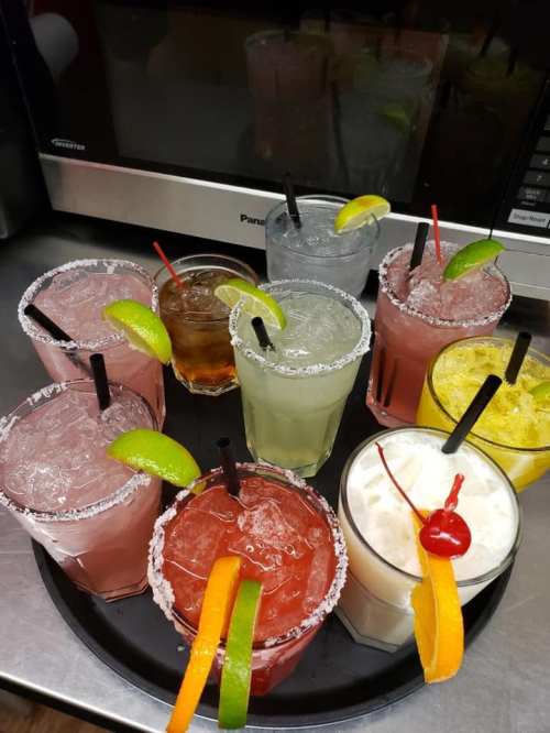 A tray of colorful cocktails with garnishes, including lime and orange slices, set against a kitchen background.