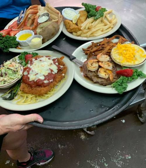 A server holds a tray with four plates of various dishes, including seafood, steak, pasta, and sides.