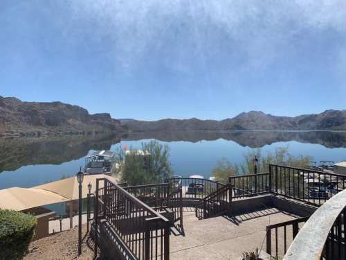 Scenic view of a calm lake surrounded by mountains, with a patio and boats in the distance under a clear blue sky.