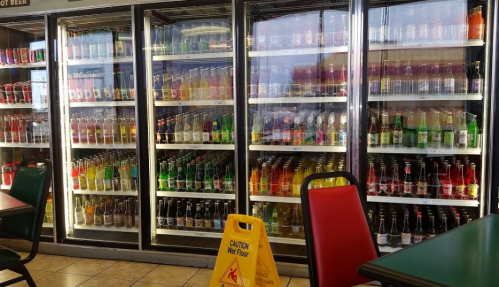 A display of various colorful soda bottles in glass refrigerators, with a caution sign on the floor.