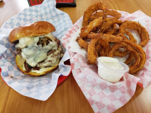 A cheesesteak sandwich with melted cheese on a bun, served with a side of crispy onion rings and dipping sauce.