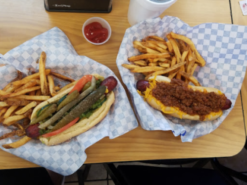 Two hot dogs on paper plates: one with toppings like pickles and tomatoes, the other with chili and cheese, served with fries.