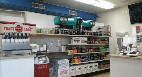 A retro-themed store interior featuring a vintage car on display, shelves stocked with various sodas, and a soda fountain.