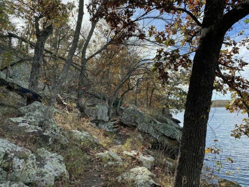 A scenic view of a rocky shoreline with trees and autumn foliage beside a calm lake under a partly cloudy sky.