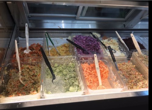 A variety of colorful salads in clear containers, with serving utensils, displayed in a refrigerated case.
