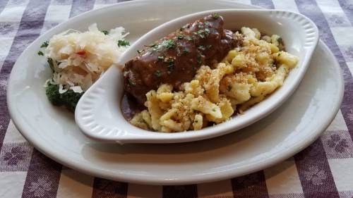 A plate featuring a sausage with gravy, spätzle, and sauerkraut, garnished with parsley on a checkered tablecloth.