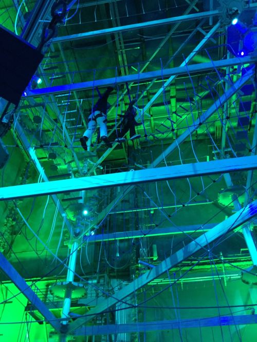 Two figures navigate a complex structure of beams and wires, illuminated by green and blue lights.