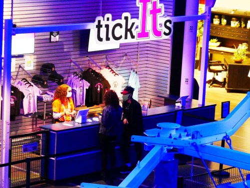 A brightly lit merchandise counter with a staff member assisting two customers in a retail space.