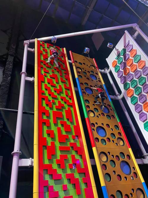Two climbers ascend colorful climbing walls with various patterns and shapes in an indoor climbing facility.