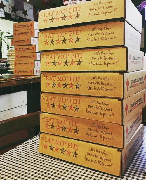 A stack of pizza boxes with the text "EAT MO' PIE!" printed on them, arranged on a checkered tablecloth.