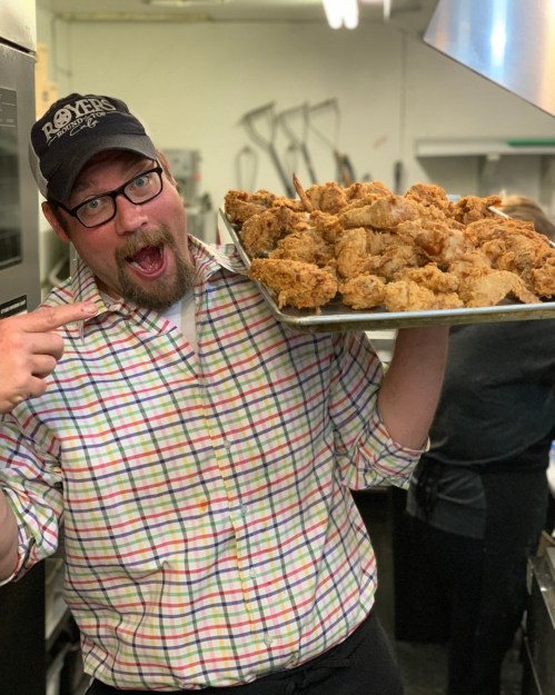 A man in a plaid shirt and cap excitedly holds a tray of golden fried chicken in a kitchen.