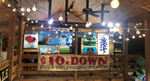 A rustic interior with string lights, featuring artwork on the walls and a sign advertising furniture for $10 down.