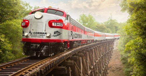A vintage red and silver train crosses a wooden trestle bridge surrounded by lush greenery.