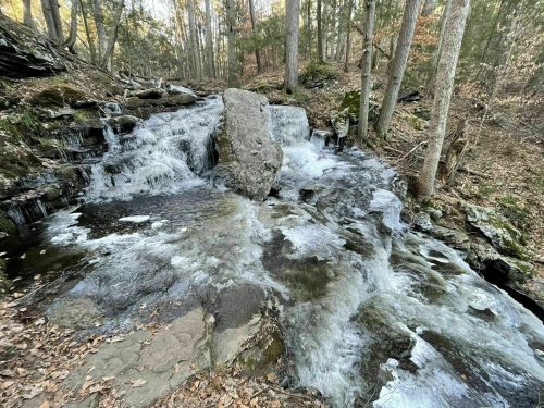 A serene waterfall cascades over rocks in a forest, surrounded by trees and autumn foliage.