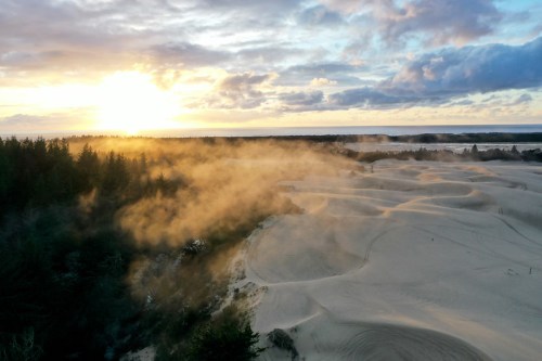 A scenic view of sand dunes at sunset, with mist rising and trees in the background under a cloudy sky.