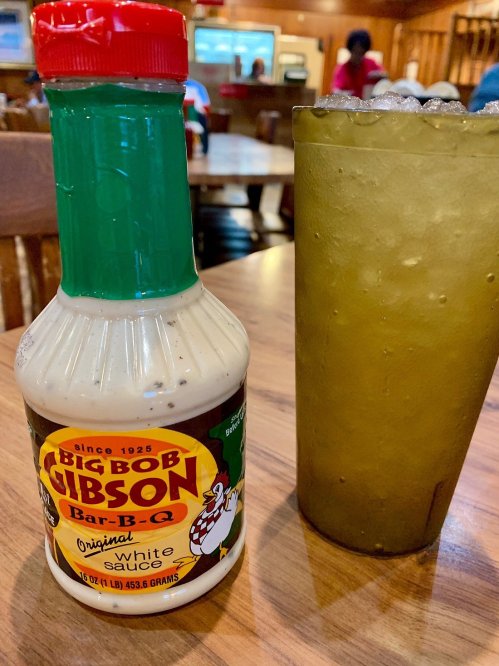 A bottle of Big Bob Gibson Bar-B-Q white sauce next to a tall glass of iced tea on a wooden table.